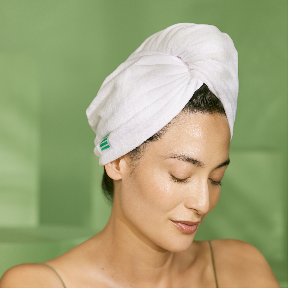 Woman with glowing skin wearing a white towel turban against a green background, representing fresh, clean skincare results.
