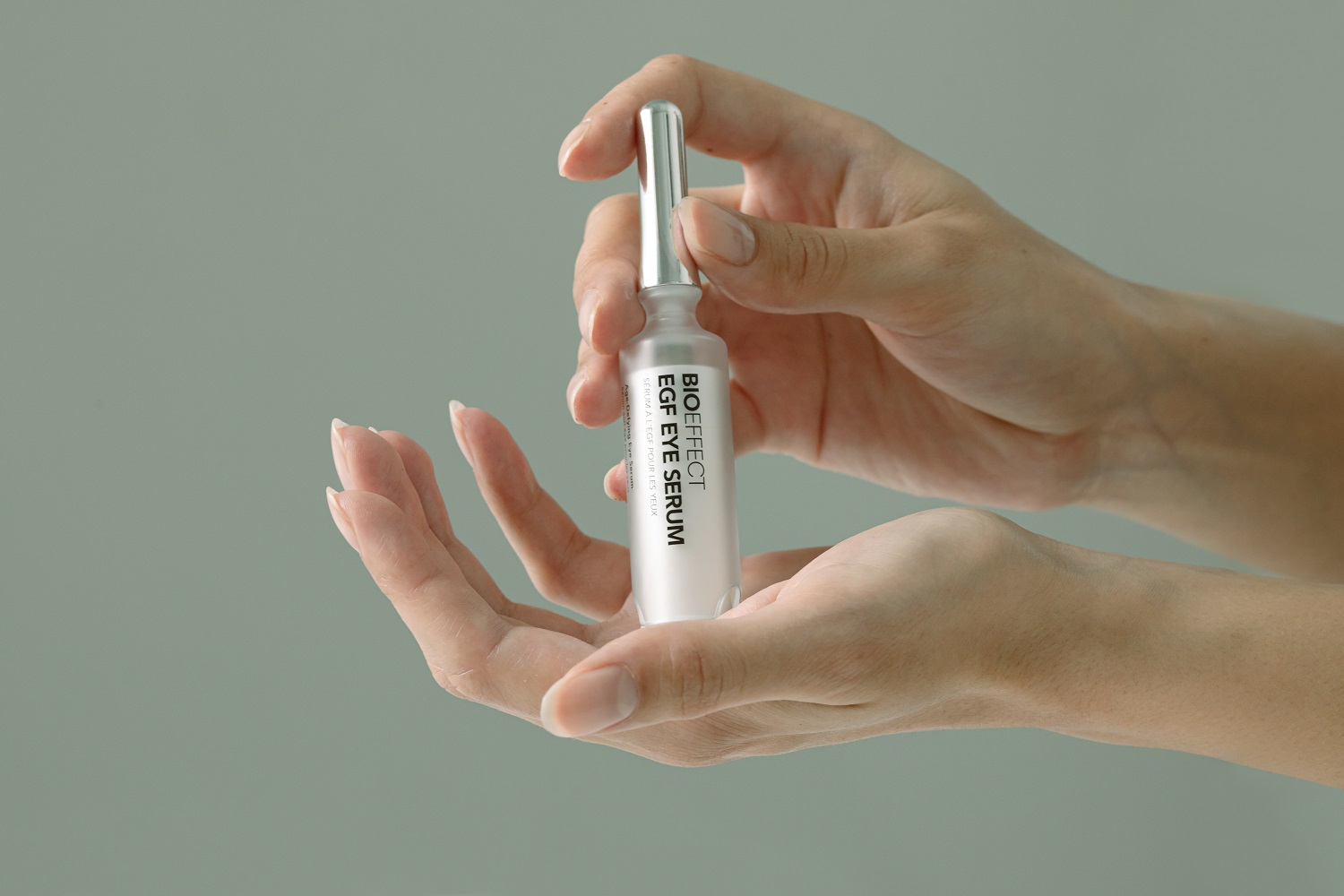 Hands holding BIOEFFECT EGF Eye Serum bottle with silver applicator against soft green background.