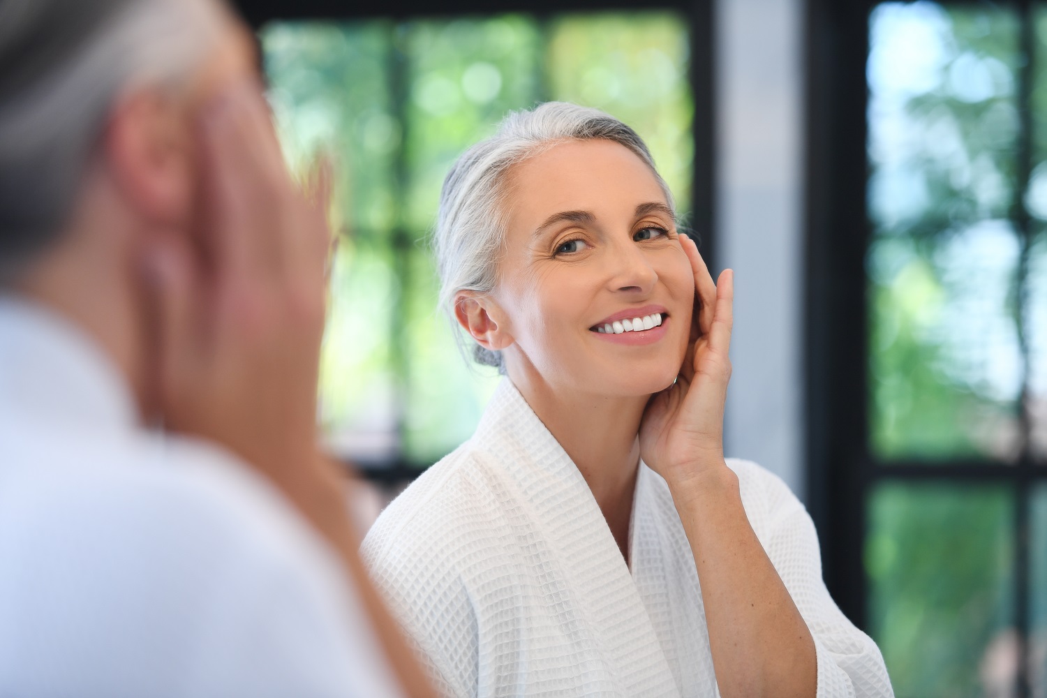 Mature woman in white robe smiling at her reflection, touching her glowing skin in a bright bathroom.