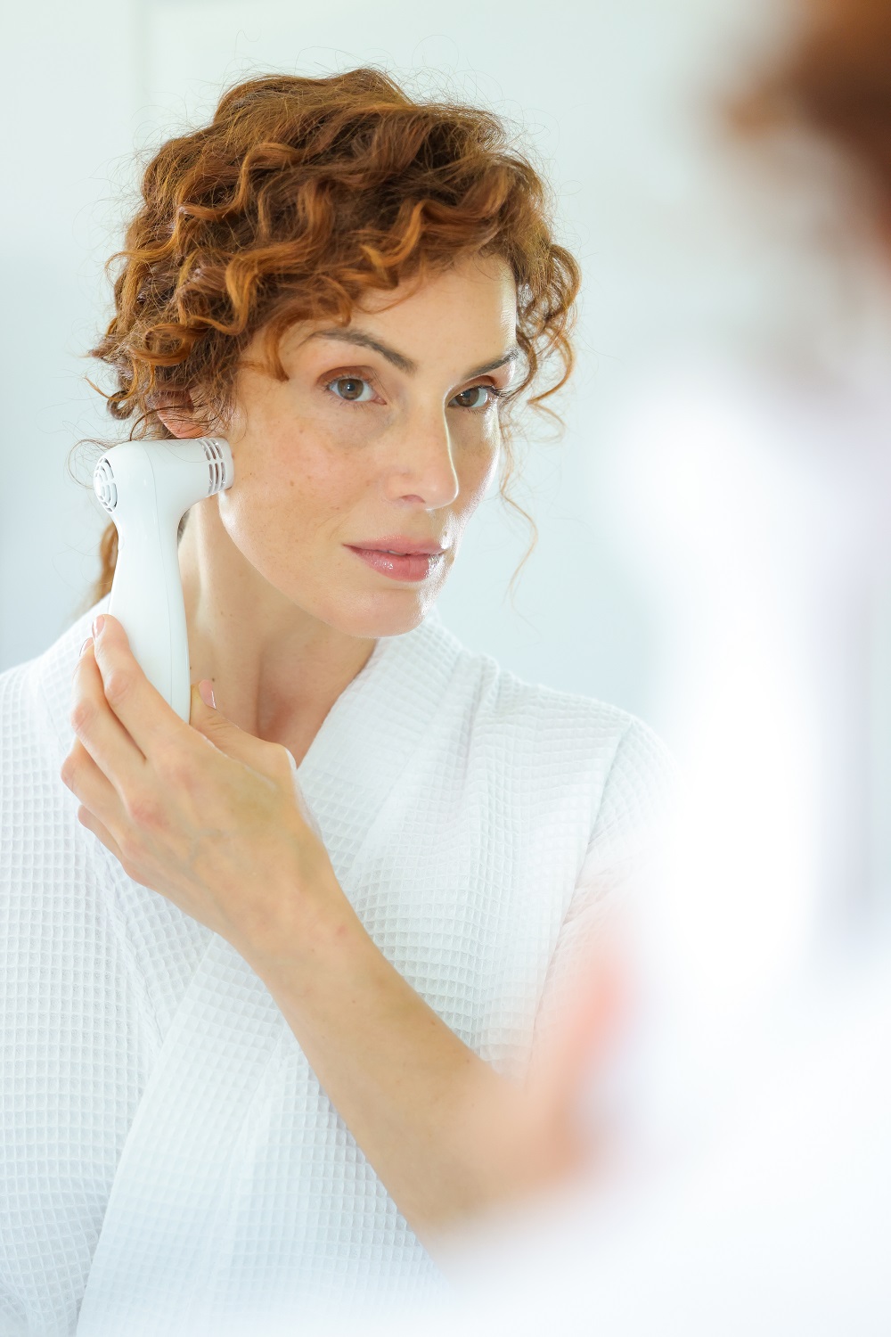 Woman in white robe using a NIRA laser device on her cheek while looking in the bathroom mirror.