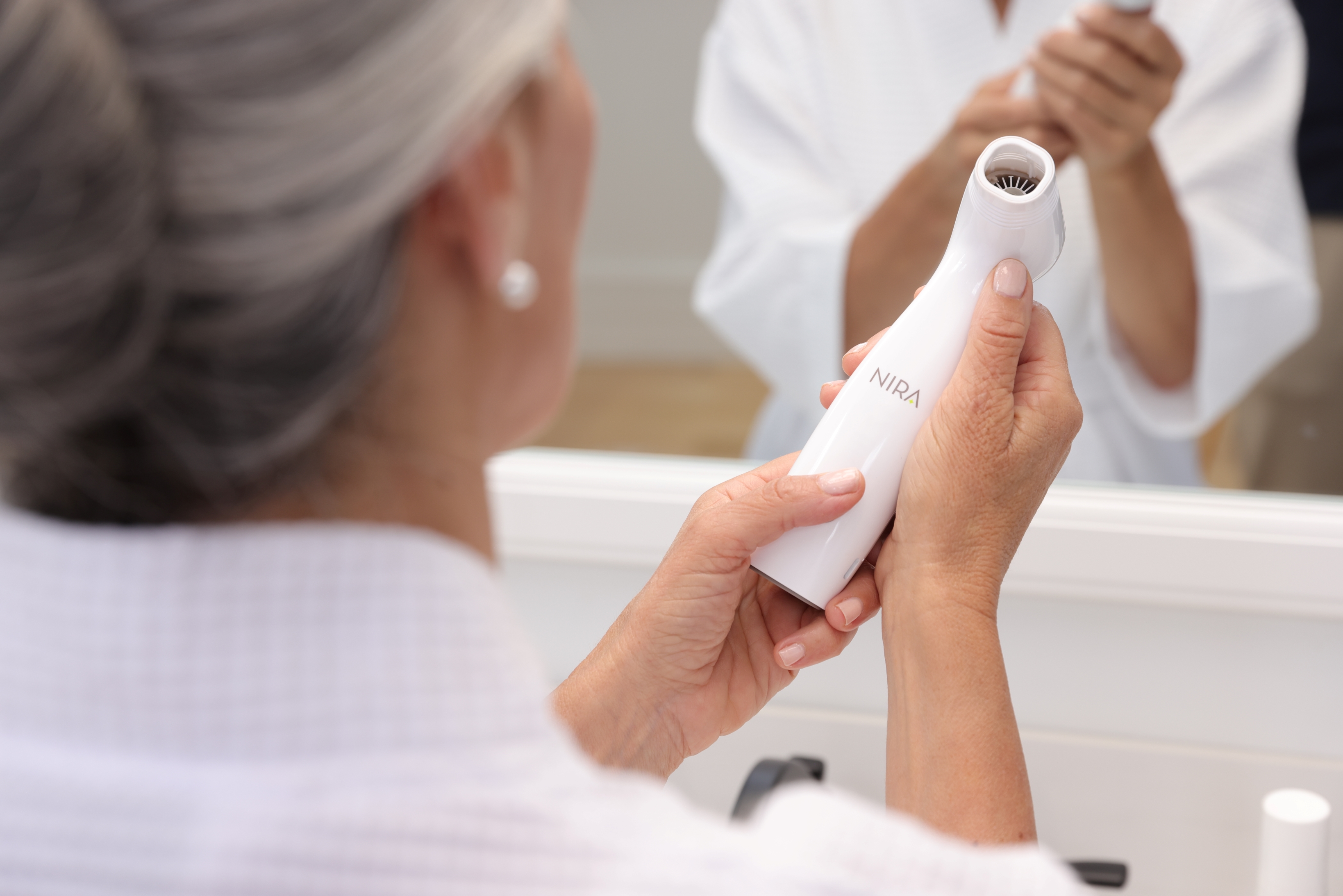 Woman in a white robe holds a NIRA laser skincare device in front of a mirror, preparing for a gentle at-home anti-aging routine.