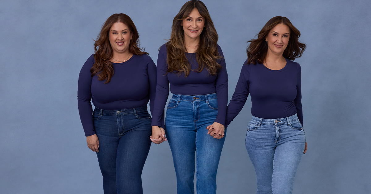 Three people in navy tops and blue jeans holding hands, standing together against a plain blue‑gray background.