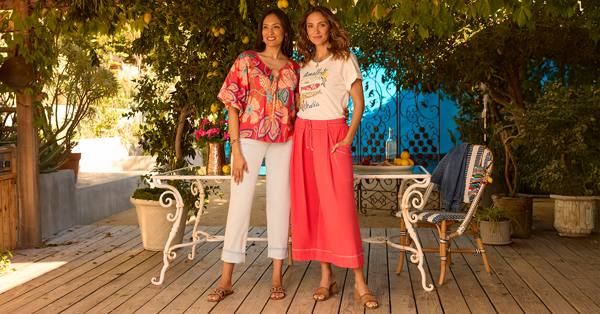 Two people in colorful summer outfits standing on a patio with flowers, lemons, and lush greenery.