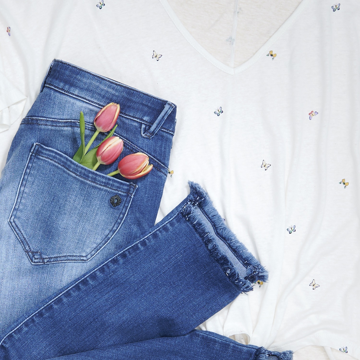 Flat lay of medium wash frayed-hem jeans with pink tulips in the pocket beside a butterfly print tee.