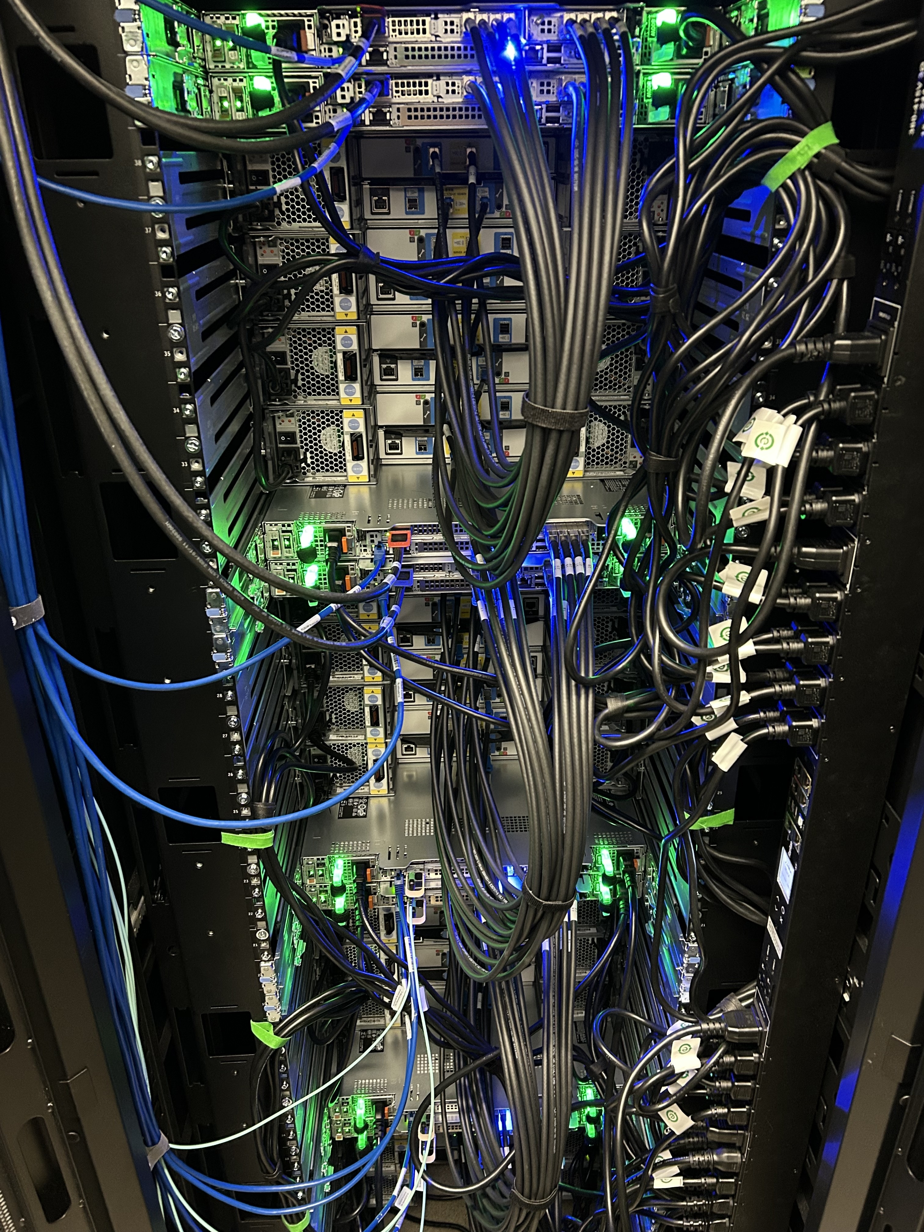 The same 5PB, from the back Rear view of a datacenter rack densely cabled with InfiniBand, SAS, and power connections between storage enclosures and servers, with green and blue LEDs illuminating the cabling.