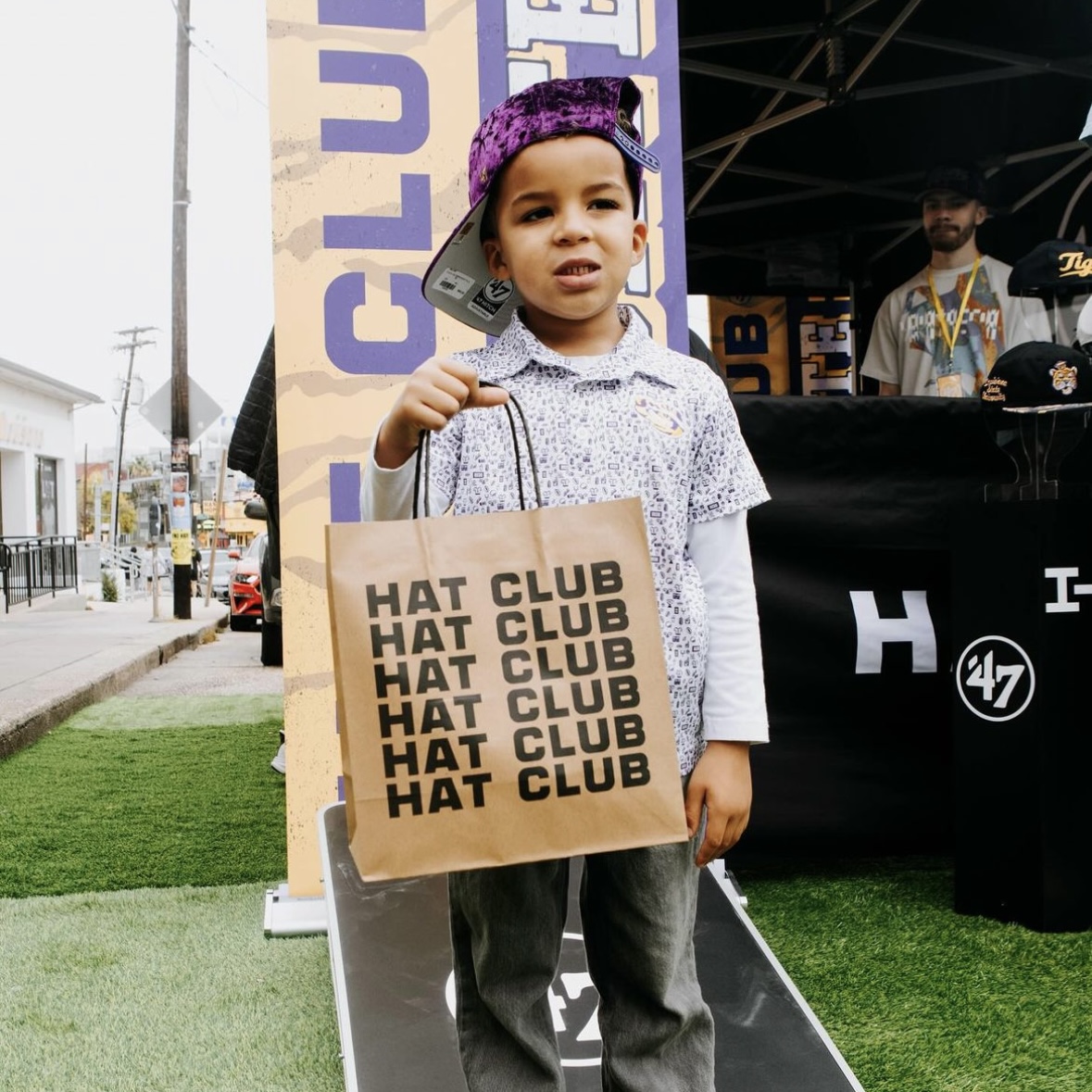 Child wearing a purple cap holds a Hat Club shopping bag at an outdoor pop-up, showcasing streetwear and MLB hat culture.
