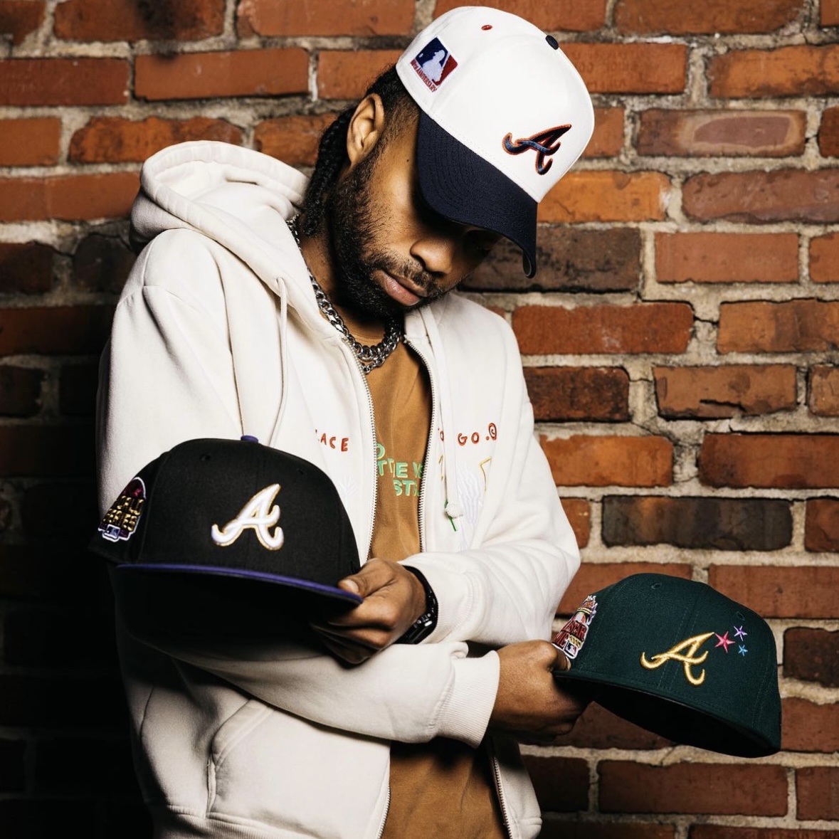Man styling Atlanta Braves fitted caps against a brick wall, showcasing Hat Club’s detailed MLB headwear and streetwear aesthetic.