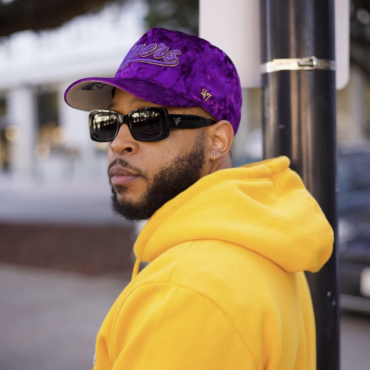 Model in purple fitted cap and yellow hoodie looks back on city street, spotlighting bold Hat Club lifestyle styling